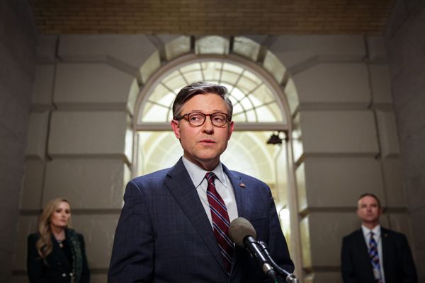 A white man with short cropped brown hair wearing a dark suit standing in front of a microphone. 