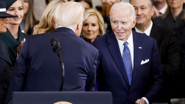 A white man in a blue suit shakes the hand of another white man wearing a blue suit. People stand around them.