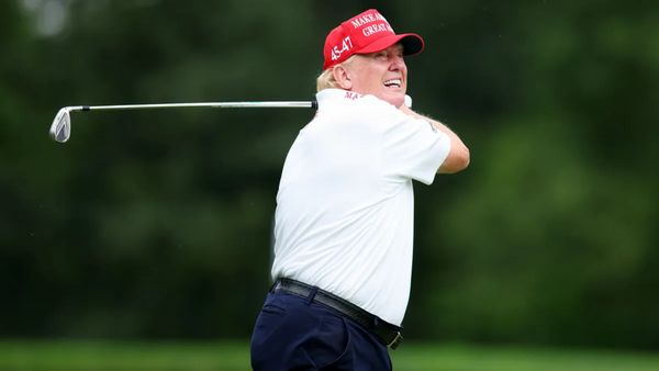 A white man wearing a white shirt and red hat that says MAKE AMERICA GREAT AGAIN in white letters smiles while holding a golf club.