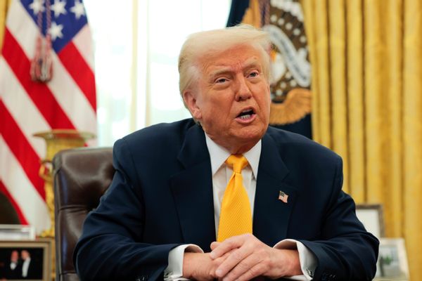 U.S. President Donald Trump, a white man with a dark tan and pale blonde hair wearing a dark suit and yellow tie, speaks with his hands crossed while seated in front of a U.S. flag and the U.S. seal. 