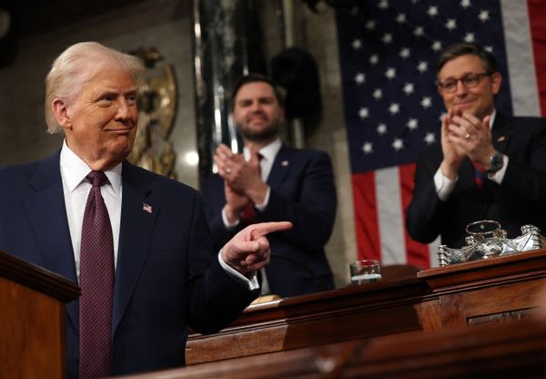 Trump, a white man with blonde hair and a dark tan wearing a suit stands behind a podium, pointing to the right. Behind him, two white men -- Vice President JD Vance and Speaker of the House Mike Johnson -- in suits stand and clap.
