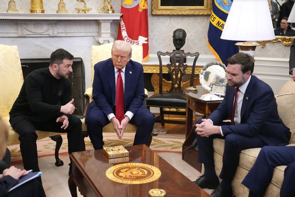 Three white men sit in a room. The one to the left is wearing a black long-sleeved shirt and pants, while the other two men are wearing dark blue suits.