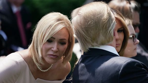 A white woman with a blonde bob wearing a white dress leans forward. A blonde man, pictured from behind, speaks into her ear. 