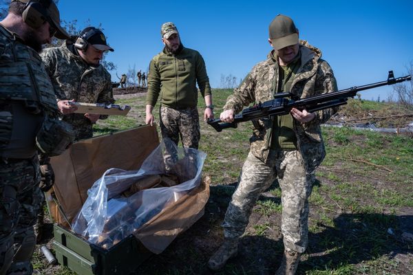 Men wearing head-to-toe camo clothing, one of whom is looking at a black machine gun. A green box with a plastic bag that presumably holds guns is next to him. 