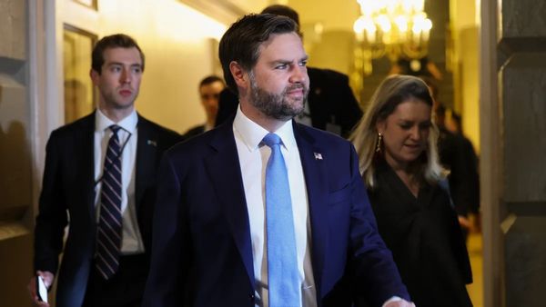 A white man in a blue suits walks looking to the right surrounded by several other people.