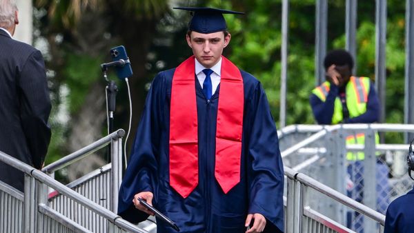 A tall white teenager in a dark graduation gown and red stole. 