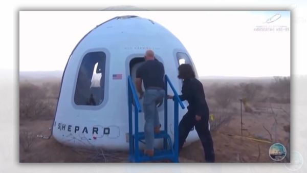 A white man stands in front of a rocket capsule's hatch. A person stands behind him.