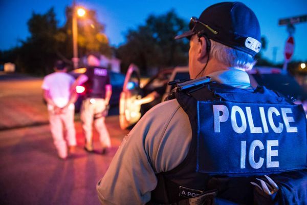 The back of an ICE officer wearing a vest which says "POLICE ICE" on it. Police lights and another official arresting someone is visible in the blurred background. 