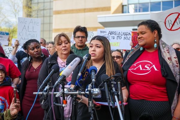 A woman with dark hair lightening to blonde speaks in front of a large number of microphones at what appears to be a news conference. She is accompanied by many other people, some of whom are holding signs protesting the deportation of Kilmar Abrego Garcia.