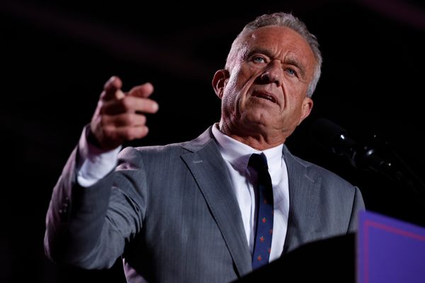 A white man with greying hair and a dark tan points at the camera. He is wearing a grey suit with a dark tie and standing behind a podium. 