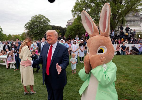 A white man holds his arms open next to a large Easter bunny. A white woman stands with her back to the camera. A large group of people is seen behind them.