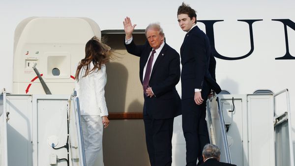 A white man turns to wave at the entrance to a plane. A white woman and man stand to his left and right respectively.