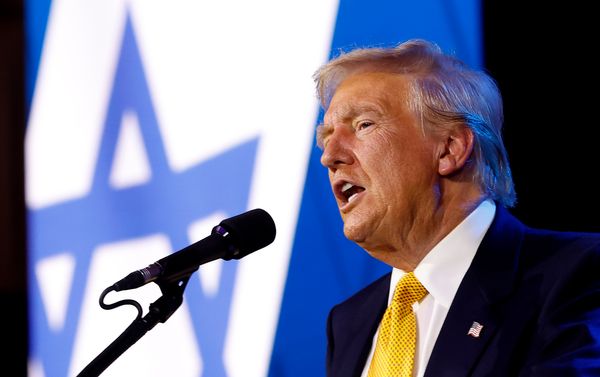 U.S. President Donald Trump, a white man with a warm tan and blonde hair wearing a suit, speaks into a microphone. Behind him is a projection of the flag of Israel, a blue and white striped flag with a Star of David in the center.