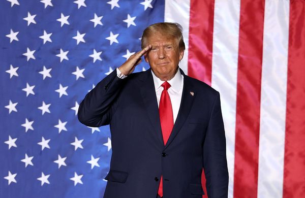 A white man wearing a dark suit salutes in front of a large red, white and blue flag.