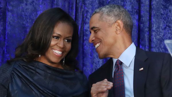 Michelle Obama, a Black woman with a bob wearing a dark dress, chats with her husband, Barack Obama, a Black man with short greying hair wearing a suit and tie.