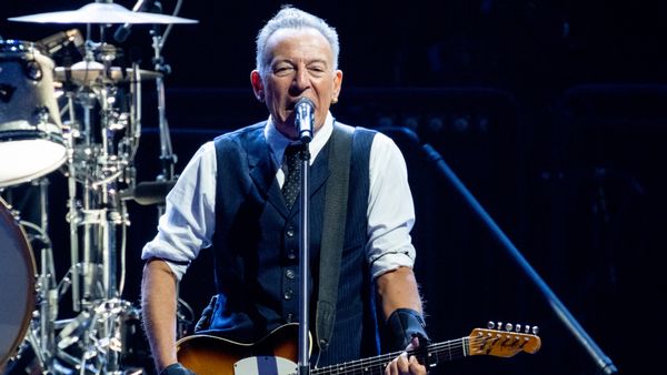 A white man stands at a microphone while playing a guitar. Part of a drumset can be seen behind him.
