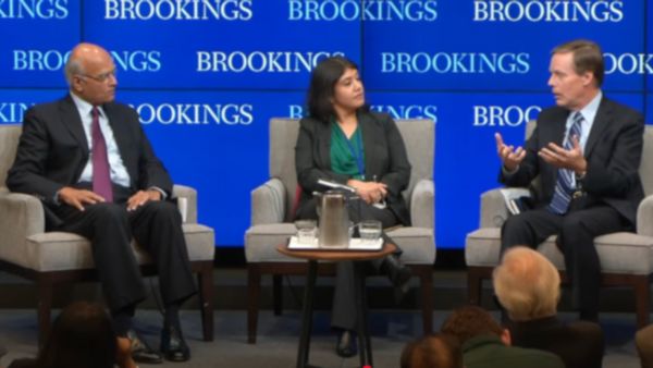 Three people sit on chairs in front of screens that say "BROOKINGS." Two of the people on the left look at the person sitting on the very right.