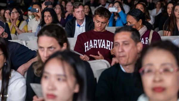 A crowd of people sit in what appears to be white folding chairs. Two people wear shirts that say HARVARD in white letters.