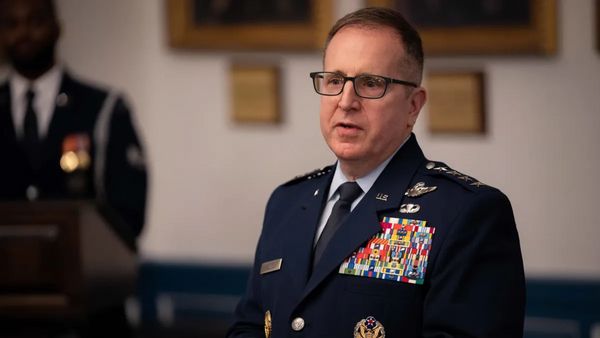 A white man wearing a blue uniform stands looking to the left. You can see another person standing in the background out of focus.