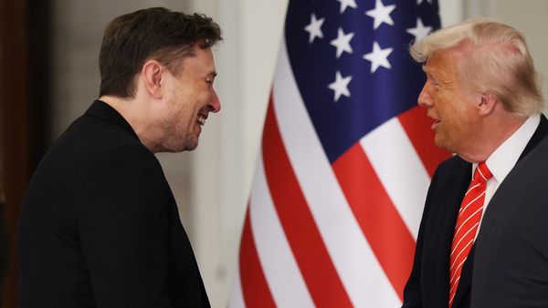Two white men wearing dark outfits smile at each other.