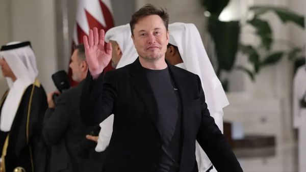 A white man wearing a dark suit jacket and shirt waves while looking at a camera. Several people stand behind him.