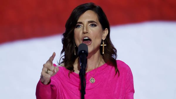 A white woman with shoulder-length hair wearing a pink shift dress points toward the camera in mid-speech.  
