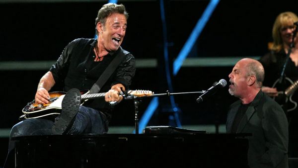 Two white men perform on stage. One holds a guitar and sits on a piano looking to his right at the person singing sitting on its bench.