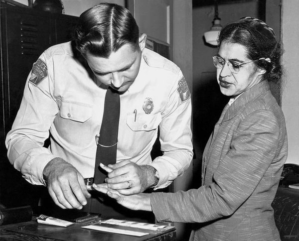 In this black and white picture, Rosa Parks, a Black middle-aged woman, gets fingerprinted during an arrest by a white police officer in a uniform. 
