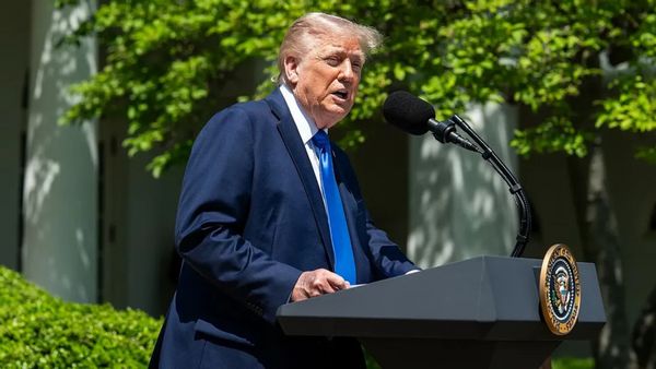 A white man wearing a dark blue suit speaks at a podium outside.