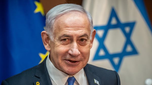 Benjamin Netanyahu, Prime Minister of Israel, in a suit stands in front of a blue and white Israeli flag.