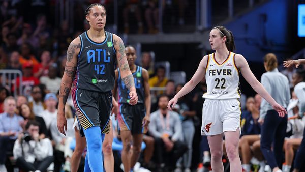 A Black women walks on a basketball court near a white woman looking to her right. A crowd of people can be seen behind them.