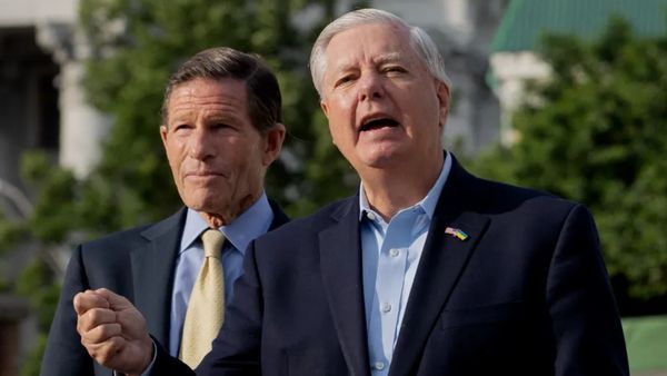 Two white men wearing dark suits stand next to each other. The one on the right appears to be speaking.