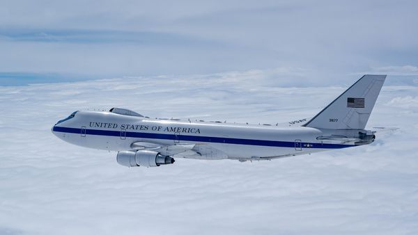 A U.S. Air Force E-4B "Nightwatch" aircraft, known as the "Doomsday Plane," flies above a blanket of clouds. The aircraft is painted white with a blue stripe and the words "United States of America" along the fuselage. The American flag and tail number 31677 are visible on the vertical stabilizer.