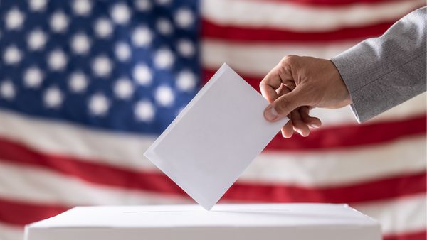 Hand holding ballot in voting ballot box with USA flag in background. 