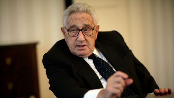 Henry Kissinger, wearing a suit, blue tie, and glasses, speaks during an interview in Washington, D.C. He is seated in a chair, gesturing with one hand, with a neutral backdrop behind him.