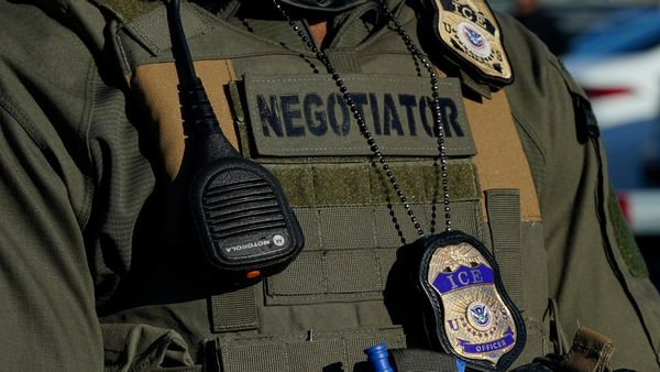 Close-up of a U.S. Immigration and Customs Enforcement (ICE) officer's tactical vest. The vest features a large patch labeled “NEGOTIATOR,” a Motorola radio microphone clipped to the front, and a metallic ICE badge hanging from a beaded chain. The officer is wearing a green uniform.