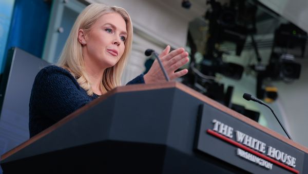 A white woman with blonde hair stands in front of a podium that says "THE WHITE HOUSE" while gesturing. 