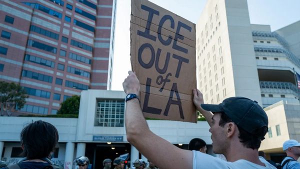 A person holds a sign that says "ICE OUT of LA." Other people can be seen around them, as well as soldiers faintly towards the bottom of the image.
