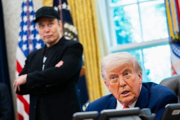 A white man wearing a blue suit is sitting down while speaking. Behind him, a white man wearing all black can be seen slightly blurred in the background.