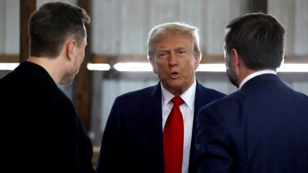 Two white men stand with their backs to the camera looking at another white man who is speaking. The one on the left appears to be wearing a black coat, while the two on the right are wearing dark blue suits.