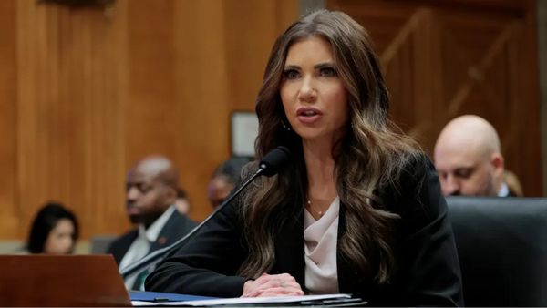 A white woman speaks into a microphone. Several people can be seen sitting behind her.