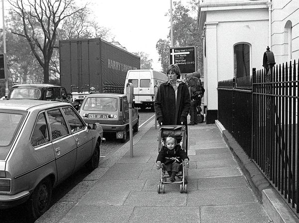 In this black and white image, a young woman pushes a stroller with a baby inside along a sidewalk. 
