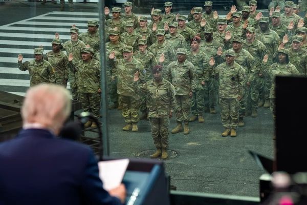 The back of a man's head in the foreground with a group of military personnel who are raising their palms and appear to be swearing an oath in the background. 