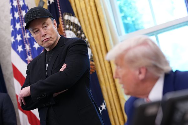 A white man wearing all black that's standing up looks down at a white man wearing a blue suit. The man wearing black is the focus of the photo.