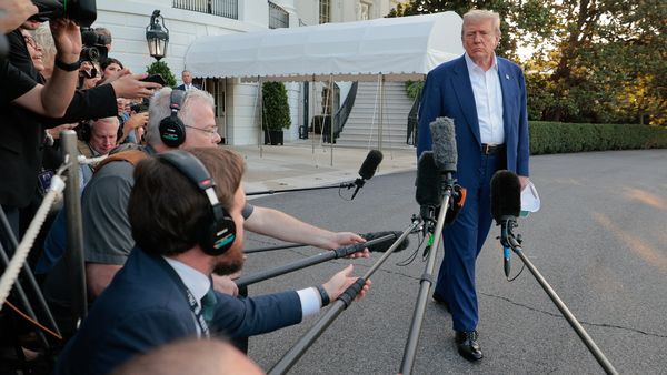 A white man stands next to a group of people. Many are wearing headphones and holding microphones in front of the man.