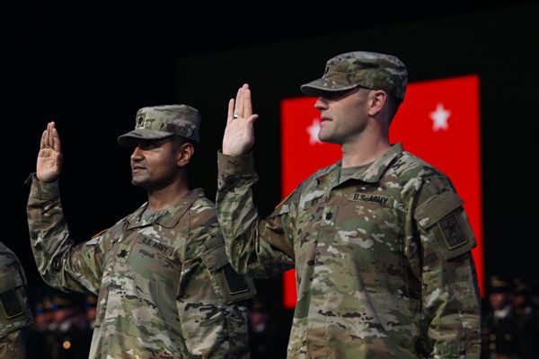 A South Asian man and a white man in camouflage military uniforms hold their palms up and appear to be swearing an oath. 
