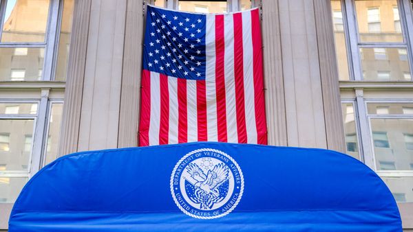 An American flag hangs vertically between columns on the VA headquarters. In front is a blue awning bearing the official seal of the U.S. Department of Veterans Affairs, featuring a bald eagle holding an American flag shield and arrows, surrounded by text and stars. 