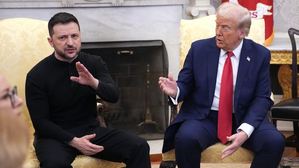 A white man wearing a black outfit sits next to a white man wearing a blue suit. The one wearing black looks forward and has his right hand raised in a slash motion, while the other man looks at him with his left hand parallel to the ground.