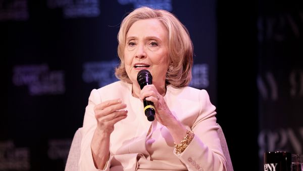 Hillary Clinton, wearing a light-colored blazer and gold bracelet, speaks into a handheld microphone during a public event. She is seated on stage in front of a dark backdrop with blurred white text.