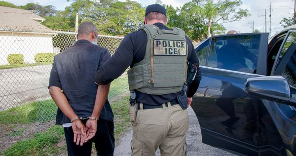 An ICE officer in a uniform holding the arm of a handcuffed person. 